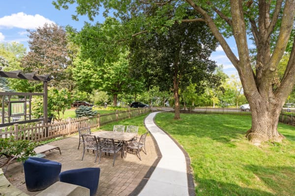 Outdoor seating area with a round table and chairs, surrounded by trees and a winding path.