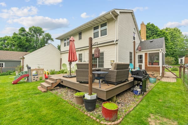 A backyard featuring a wooden deck with wicker seating, a gas grill, and a children's play structure.