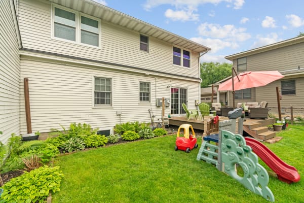 Backyard of a two-story home featuring a lawn, a toy car, playground equipment, and a shaded deck.