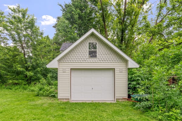 A small garage with a white door and scalloped shingles set in a green landscape.