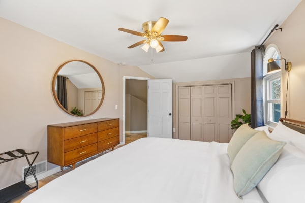 A well-decorated bedroom with a white bed, wooden dresser, large mirror, and modern ceiling fan.