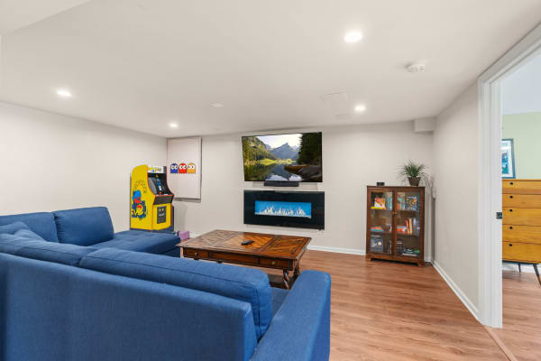 A modern basement living room with a blue sectional sofa, arcade game, flat-screen TV, and electric fireplace.