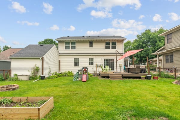 Backyard view featuring a house, deck with furniture, children's play slide, and garden areas.