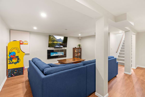 Modern basement living space with blue sofa, electric fireplace, TV, and arcade game.