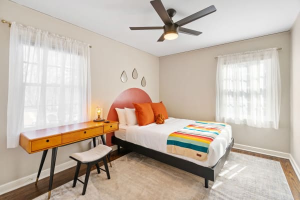 Modern bedroom with a bed, wooden desk, and decorative items, illuminated by natural light.