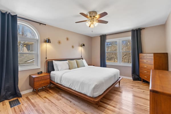 A modern bedroom featuring a white bed, wooden furniture, and large windows with dark curtains.