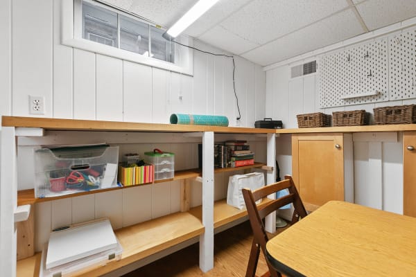 A well-lit basement workspace featuring a wooden workbench, storage containers, and a small table.