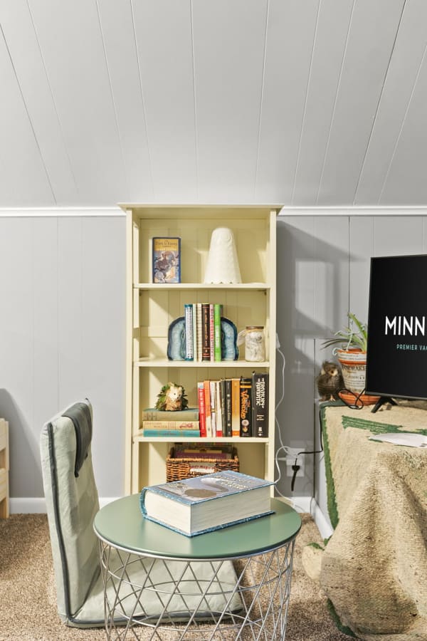 A cozy reading corner featuring a yellow bookshelf filled with books and decorative items, a green accent table with a large book, and a stylish gray chair.