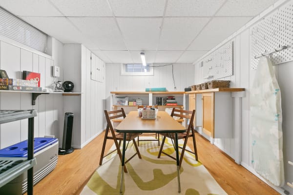 A basement interior featuring a wooden table surrounded by four chairs, board games on shelves, and light wooden flooring.