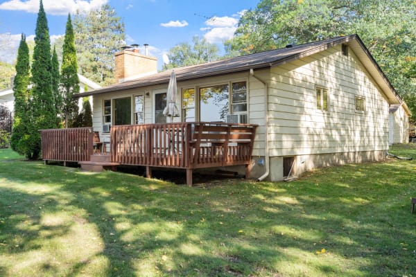 Single-story house with a wooden deck and evergreen trees in a grassy yard.