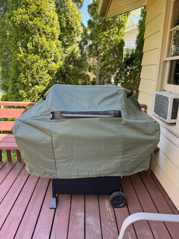 A grill covered with an olive-green cover on a wooden deck, with trees in the background.