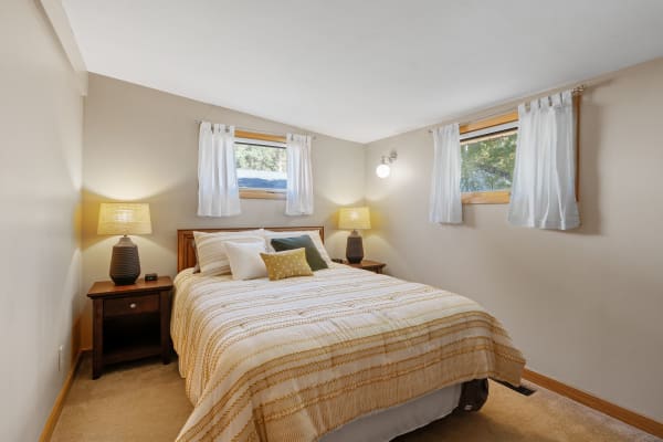 A cozy bedroom featuring a bed with decorative pillows, a bedside table with a lamp, and natural light coming from windows.