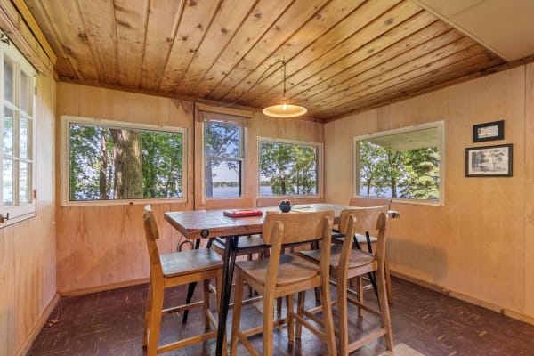 Rustic cabin interior with wooden dining furniture and windows overlooking trees and water.