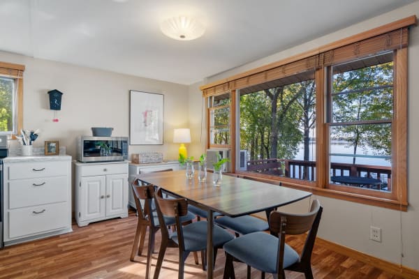 A bright dining space featuring a wooden table, chairs, and large windows overlooking a lake.