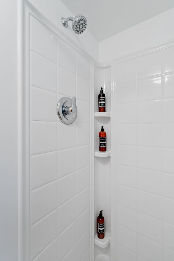 A shower with a chrome showerhead and dark amber bottles on a white tiled wall.