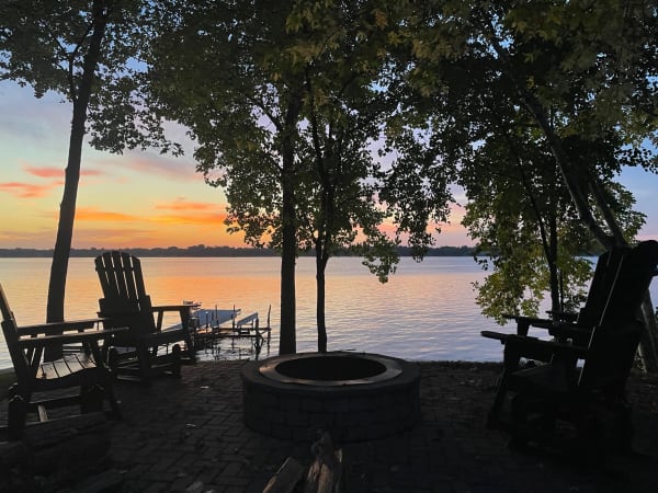 Lakeside view at sunset with a fire pit and wooden chairs under trees.