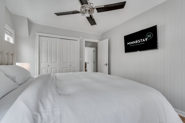 A well-lit modern bedroom with white bedding, a lamp, and a TV mounted on the wall.