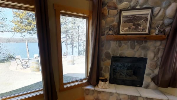 Interior view of a cozy living room with a stone fireplace and windows overlooking the lake with outdoor seating.