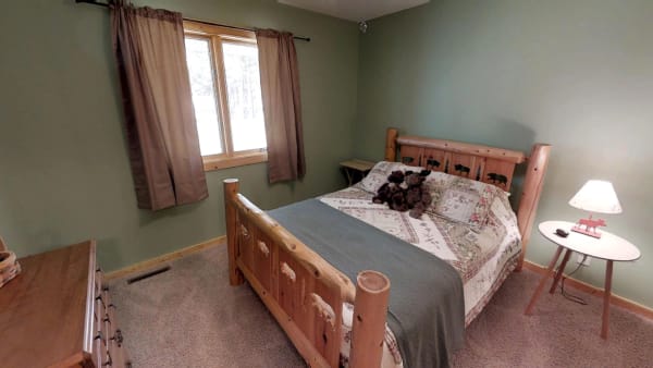 Cozy bedroom featuring a wooden bed, patterned quilt, and soft carpet with natural light from a window.