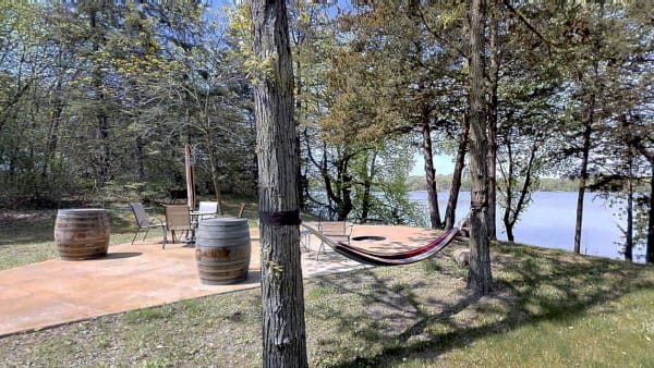 Lakeside patio featuring a hammock, wooden barrels, and a table with chairs surrounded by trees.