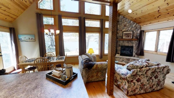 Interior of a cabin featuring a spacious living area with floral couches and a dining table, large windows overlooking a lake.