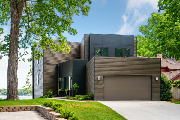 Contemporary house with dark exterior panels, large windows, and a landscaped yard overlooking a lake.