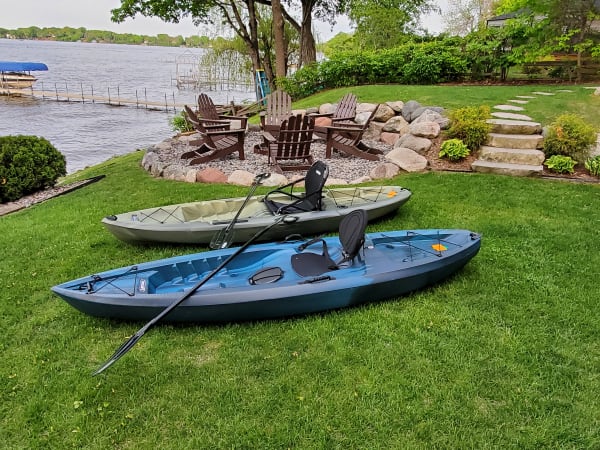 Two kayaks sit on a green lawn beside a lake, with Adirondack chairs nearby.