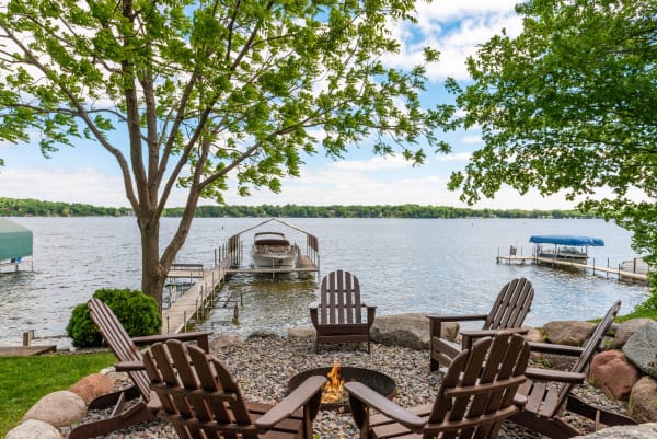 A peaceful lakeside scene with wooden chairs around a fire pit and a dock with boats.