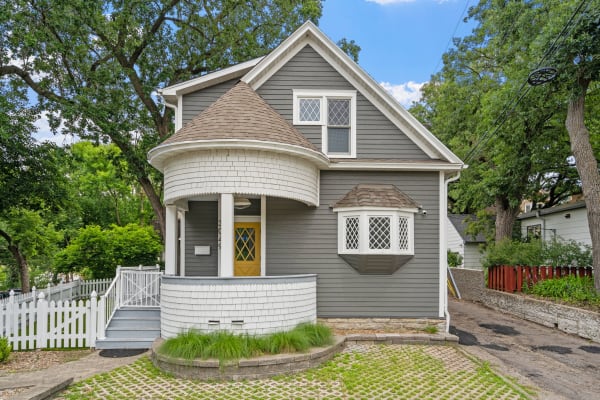 A grey house featuring a round turret entrance and yellow door, surrounded by greenery and a white picket fence.
