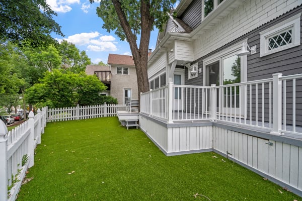 A suburban yard with artificial grass surrounded by a white picket fence and a charming house.