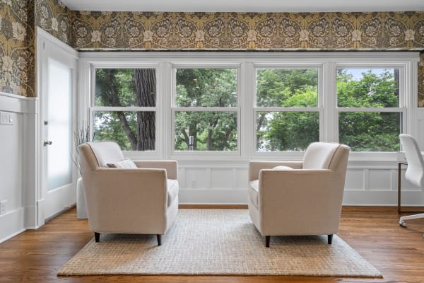 Two beige armchairs facing each other in a bright room with green trees visible through large windows.