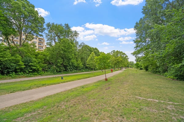 A park pathway lined with trees, an apartment building in the background, and people walking in the distance.