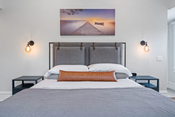A serene bedroom with a grey bed, sunset landscape photo, and stylish bedside lamps.