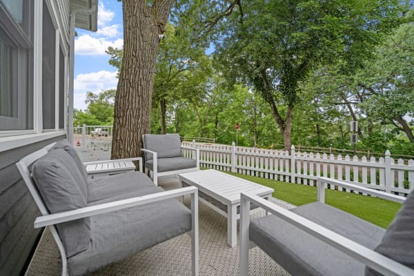 An outdoor seating area with gray chairs and a white table, framed by trees and a picket fence.