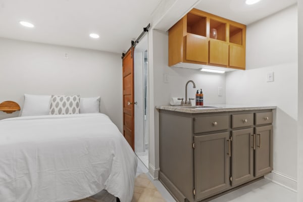 A small room featuring a bed, decorative pillow, and kitchenette with shelves.
