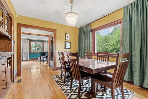 A cozy dining room featuring a wooden table, chairs, and a chandelier, with large windows and a view of greenery.