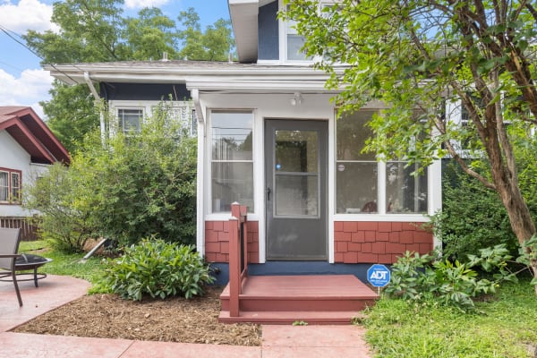 A two-story house with a red wooden porch, grey door, and surrounded by green bushes and plants.