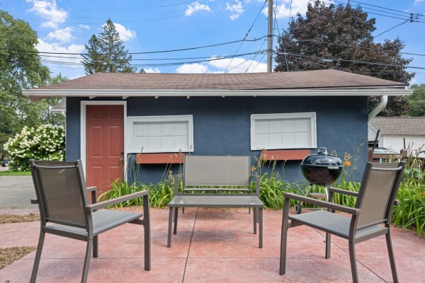 Outdoor seating area with gray chairs, a small table, and a grill in front of a blue house.
