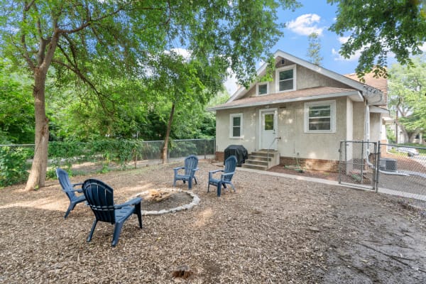 A backyard with a house, fire pit, and Adirondack chairs under green trees.