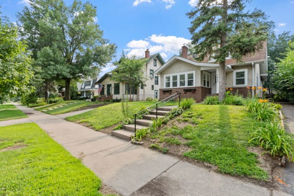 Two houses in a residential area with green lawns and blooming flowers under a blue sky.