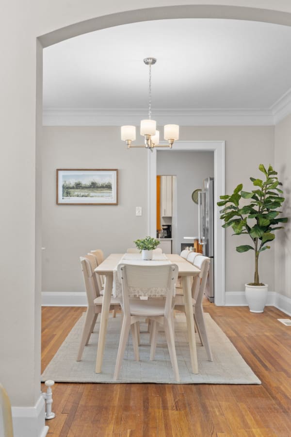 A dining room with a wooden table, chairs, plants, and an art piece on the wall.