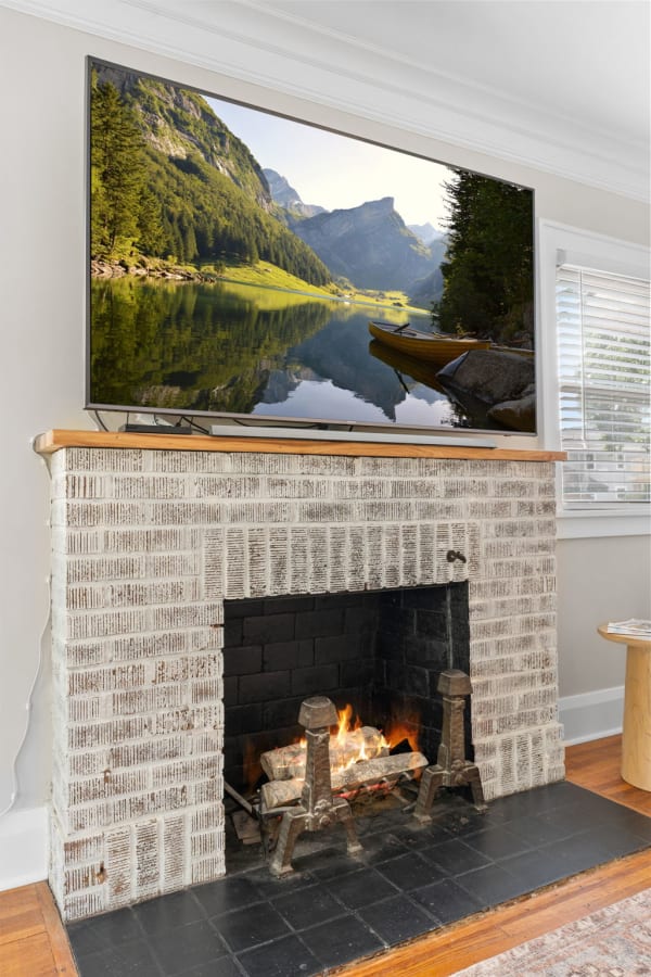 A living room featuring a brick fireplace with a fire burning, and a large TV showing a mountain lake scene.