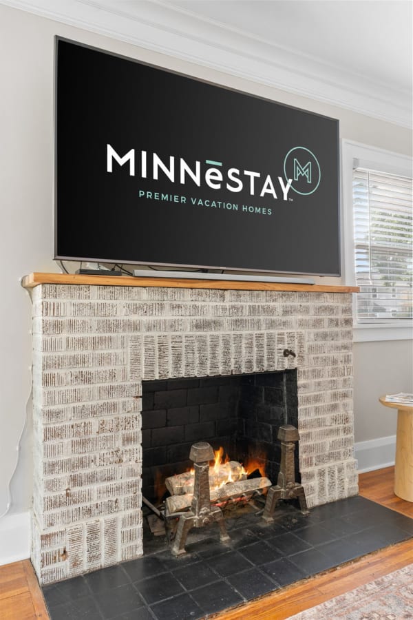 Living room with a flat-screen TV showing MINNESTAY logo above a fireplace with a flickering fire.