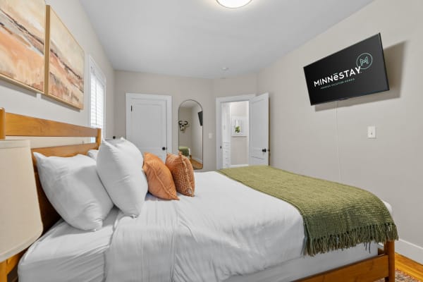 Cozy bedroom with white bedding, accent pillows, green throw, abstract art, and a wall-mounted TV.