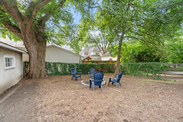 Backyard with blue Adirondack chairs around a fire pit, surrounded by trees and greenery.