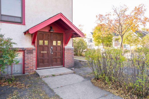 A cozy building entrance with double doors under a red overhang, surrounded by autumn foliage.