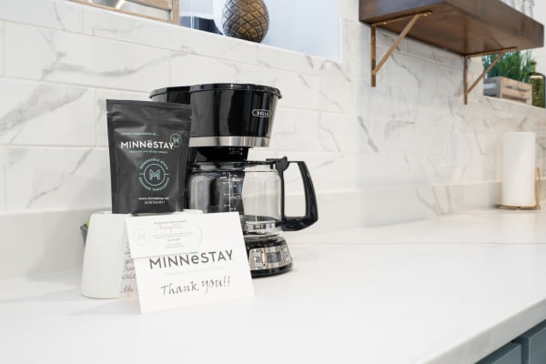 A black coffee maker on a white countertop next to a bag of MINNESTAY coffee and a thank-you card.