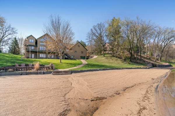 Lakeside property with a sandy beach, stone wall, and a two-story house on a green lawn.