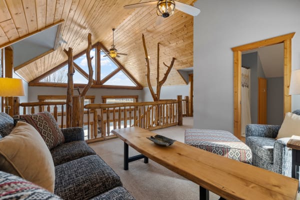 A rustic loft interior featuring a wooden ceiling, comfortable sofa, and large windows.
