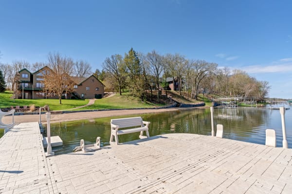 Lakeside dock with a bench, homes, and clear blue sky.